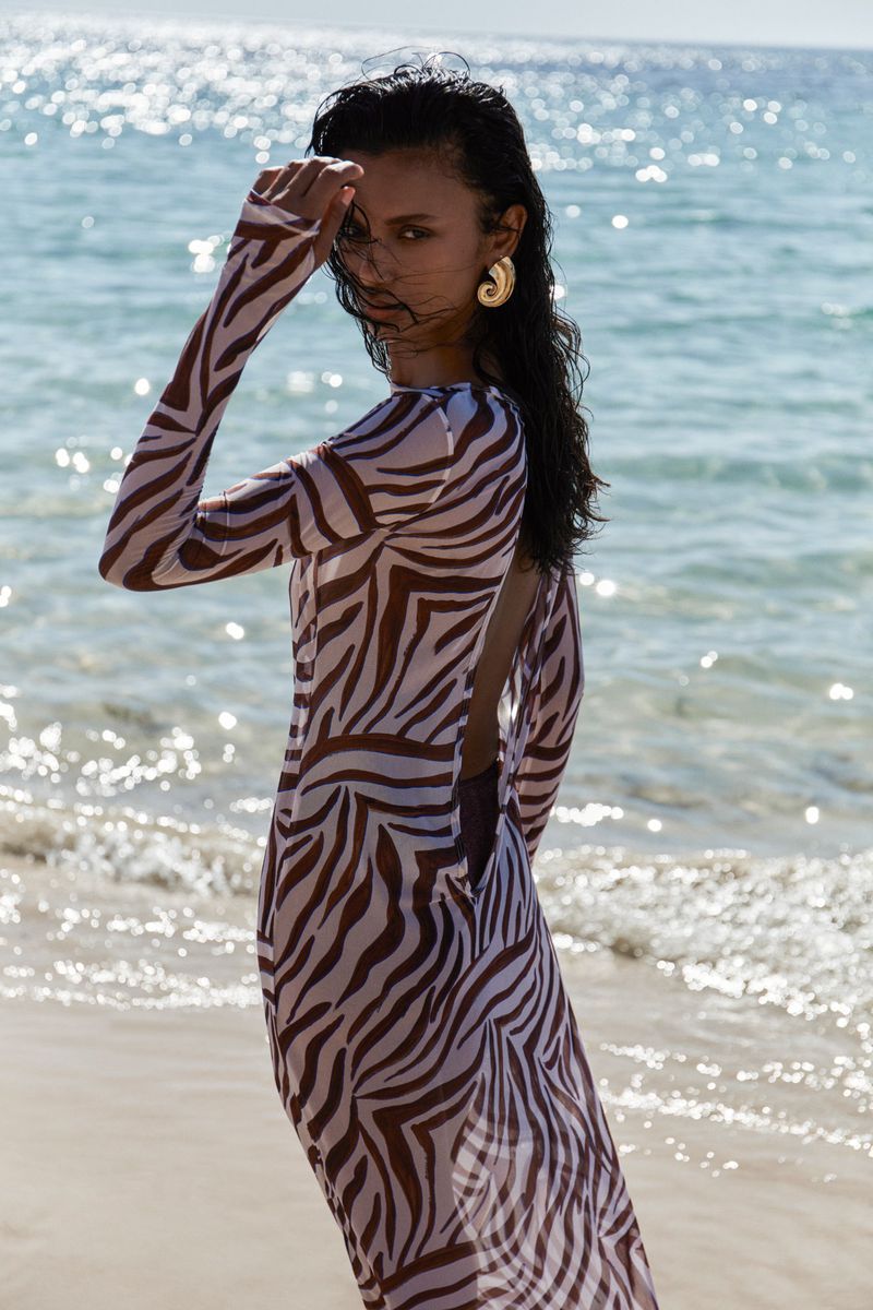 A woman stands on a sandy beach near the ocean, wearing a brown and white zebra-print dress and gold earrings in outdoor setting.