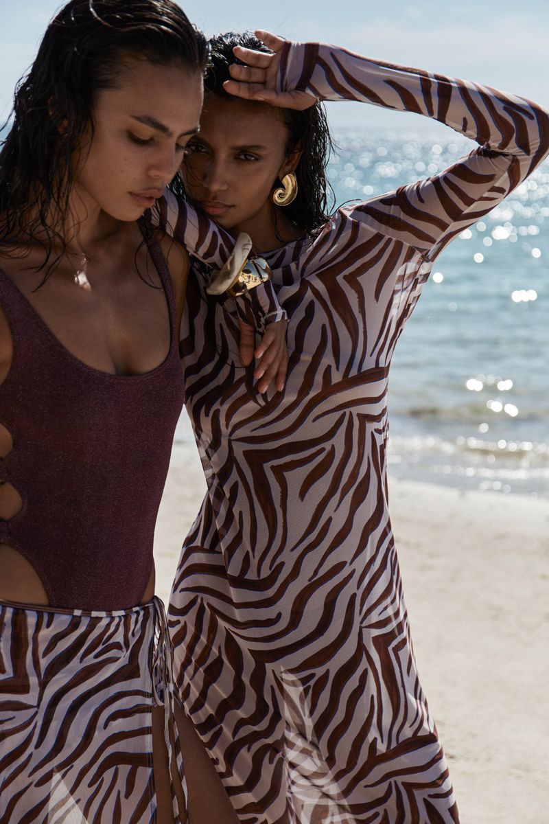 Two women pose on a sandy beach with a bright, sunlit ocean backdrop. One wears a zebra-print dress; the other, a shimmering plum swimsuit.