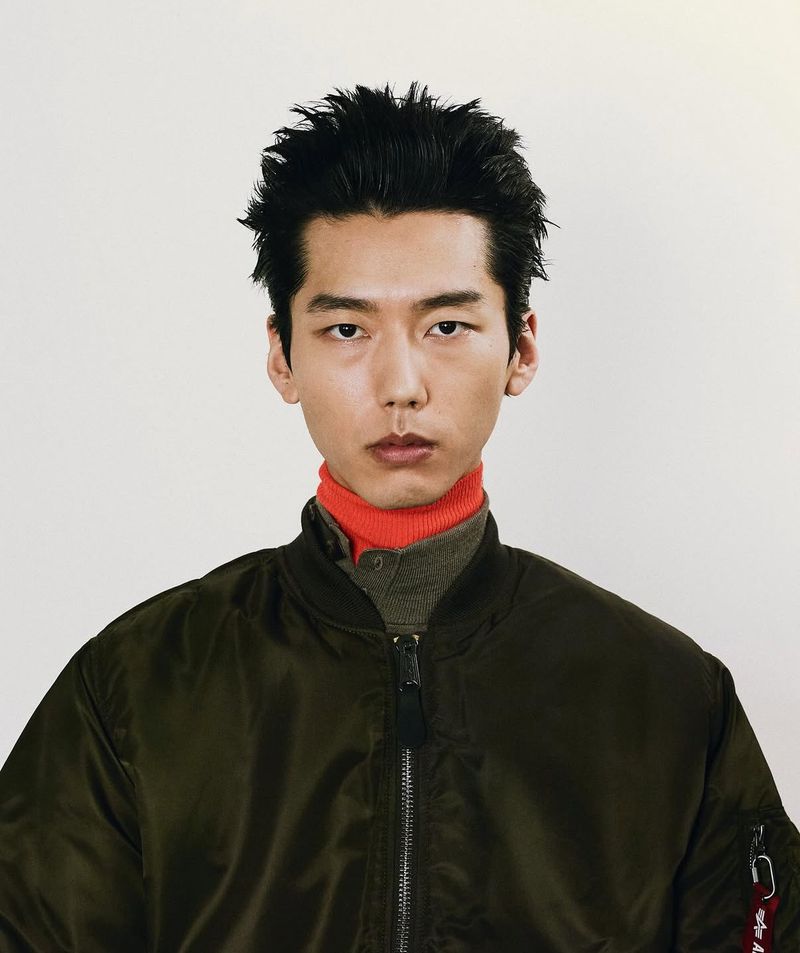 Close-up portrait of a young adult man with dark hair wearing a dark green bomber jacket with a red turtleneck against white background.