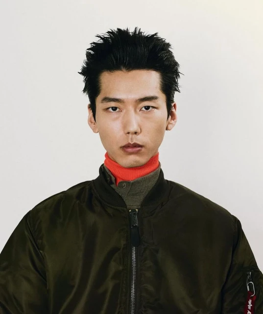 Close-up portrait of a young adult man with dark hair wearing a dark green bomber jacket with a red turtleneck against white background.