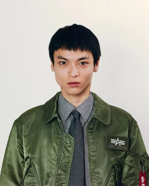 Portrait of a young Asian adult male with dark hair wearing an olive green bomber jacket, grey dress shirt, and dark grey tie against a white background.