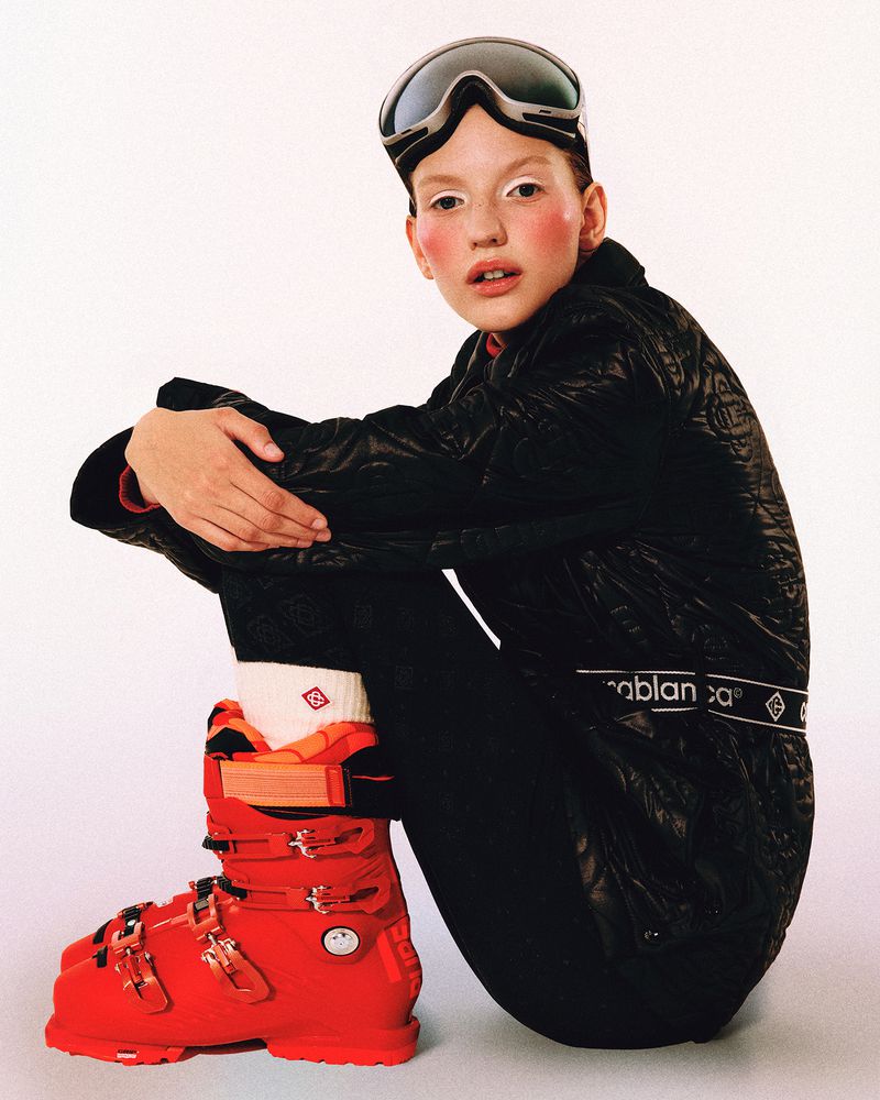 Fashion shot of a child posing while wearing a black jacket, ski goggles and boots against an off-white background in a studio.