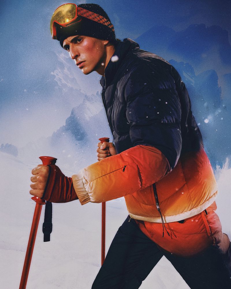 A man in ski gear, including a puffer jacket, ski goggles, and ski poles, poses against a blurred snowy mountain background.