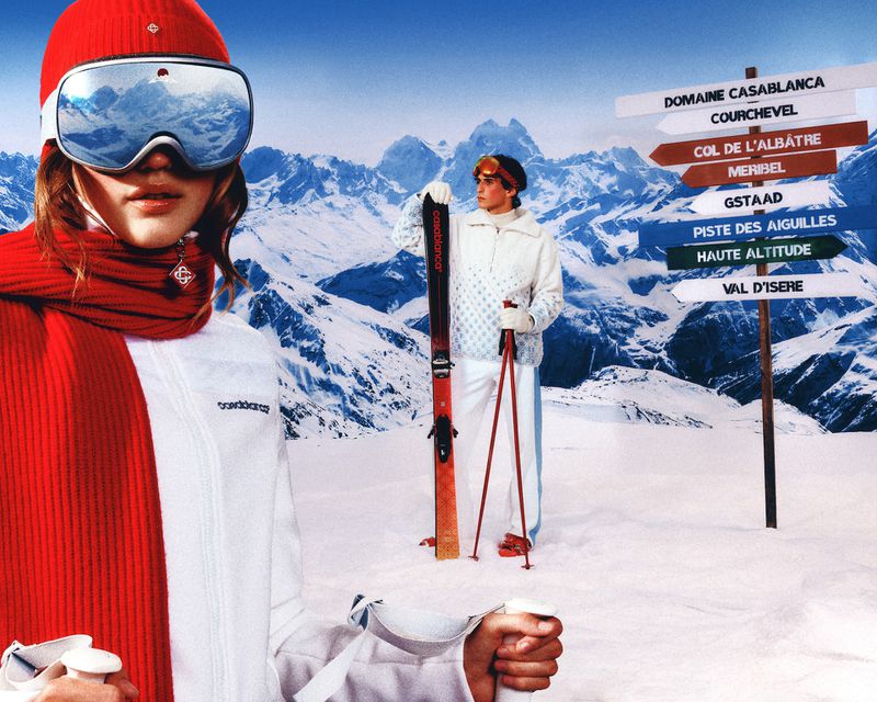Two people in ski attire pose in a snowy mountain landscape with ski equipment and a directional sign displaying destination names.