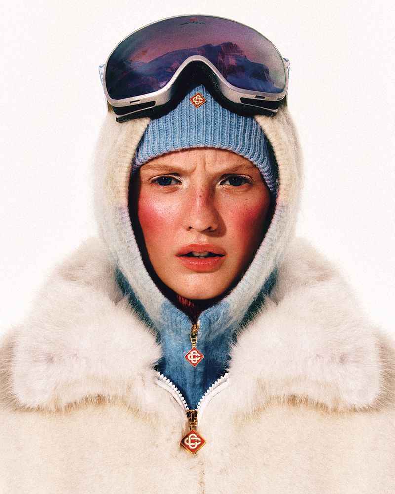 Close-up studio shot of an adult female model wearing ski goggles, a light blue beanie, and a hooded jacket with fur trim.