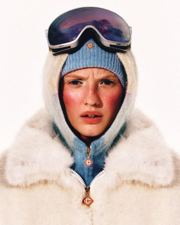 Close-up studio shot of an adult female model wearing ski goggles, a light blue beanie, and a hooded jacket with fur trim.