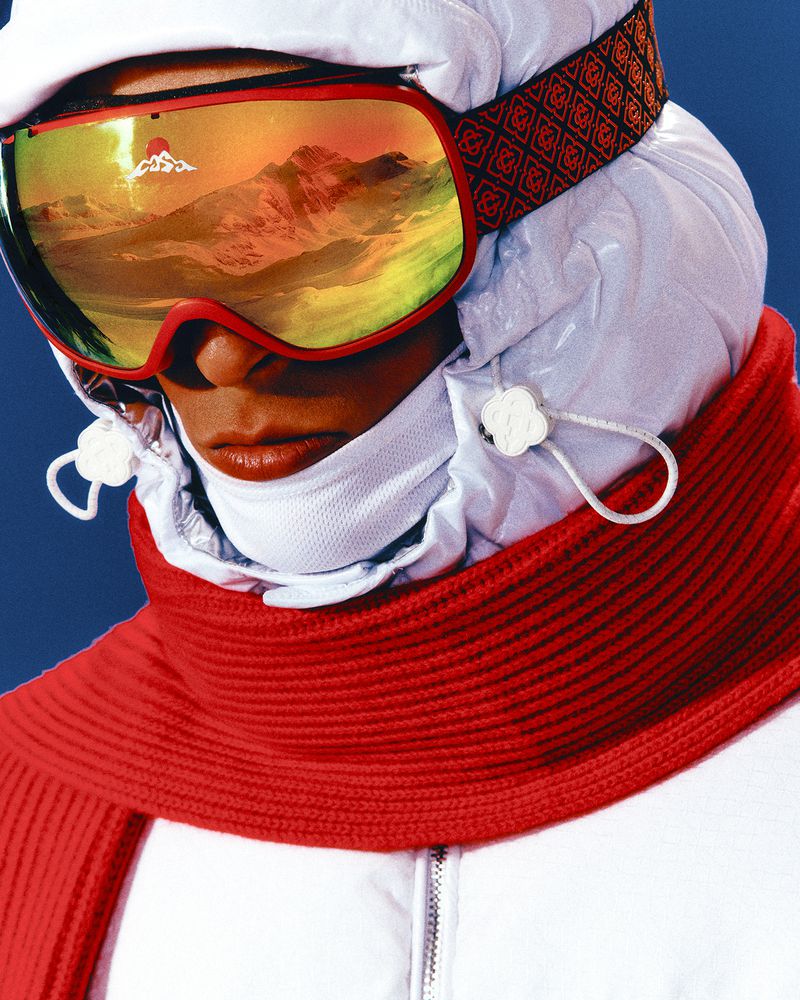 Close-up of a person in ski goggles reflecting a mountain landscape, wearing a white ski hood and red scarf against a blue backdrop.