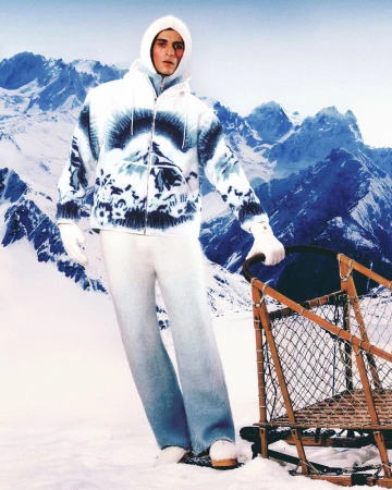 Male model in a white patterned hooded jacket and light blue jeans stands next to a wooden sled in a snowy mountain landscape.