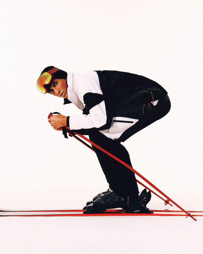 Man in ski suit and goggles in studio. He is bent over with ski poles, poised to ski. He wears a black and white ski suit.