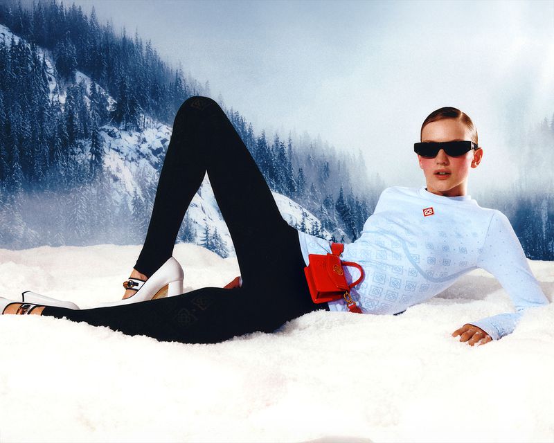 Fashion shot of a woman in a light blue patterned top and black pants, posing in the snow with a snowy mountain background.