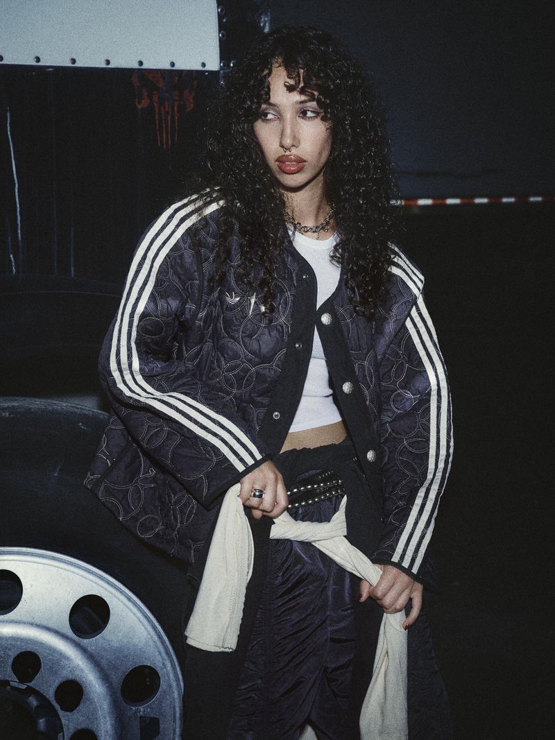 An adult female with curly hair and a patterned jacket is posing next to a truck tire in an indoor setting with dark lighting.