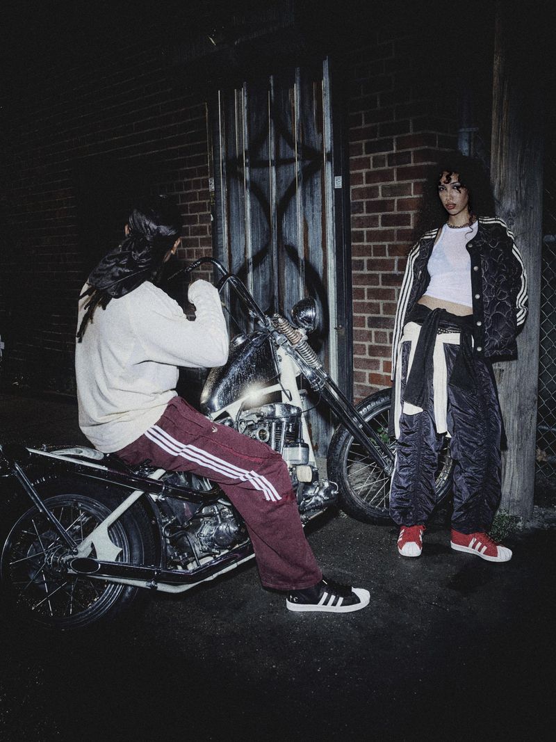 Two people in streetwear posing with a motorcycle at night in front of a brick wall and metal door, showcasing urban fashion.