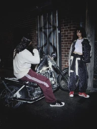 Man seated on a motorcycle next to a standing woman wearing Adidas apparel against a brick wall at night.