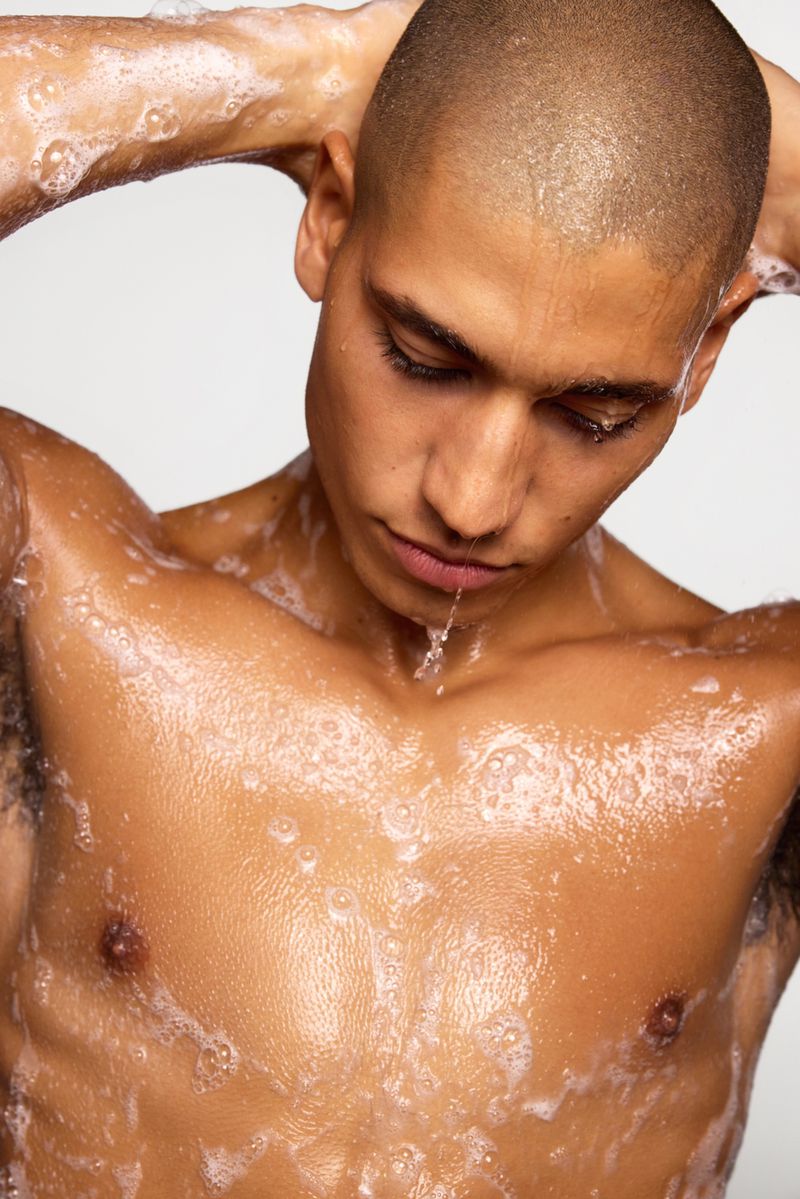 Close-up of a shirtless man with a shaved head, covered in water and soap suds, looking down with his hands behind his head.