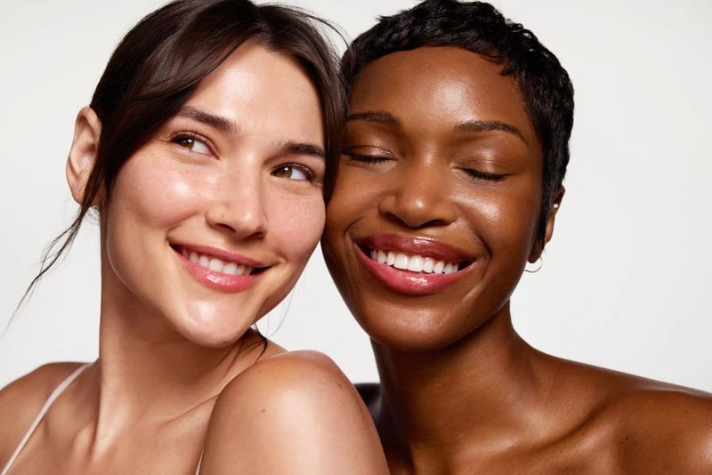 Close-up of two smiling women with radiant skin. One has dark hair and is looking to the left. The other has short hair with eyes closed.