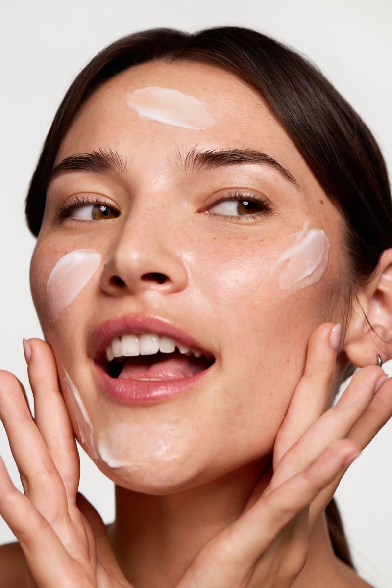 Portrait of a young woman with fair skin applying moisturizer to her face in a beauty skincare product advertisement on a white background.