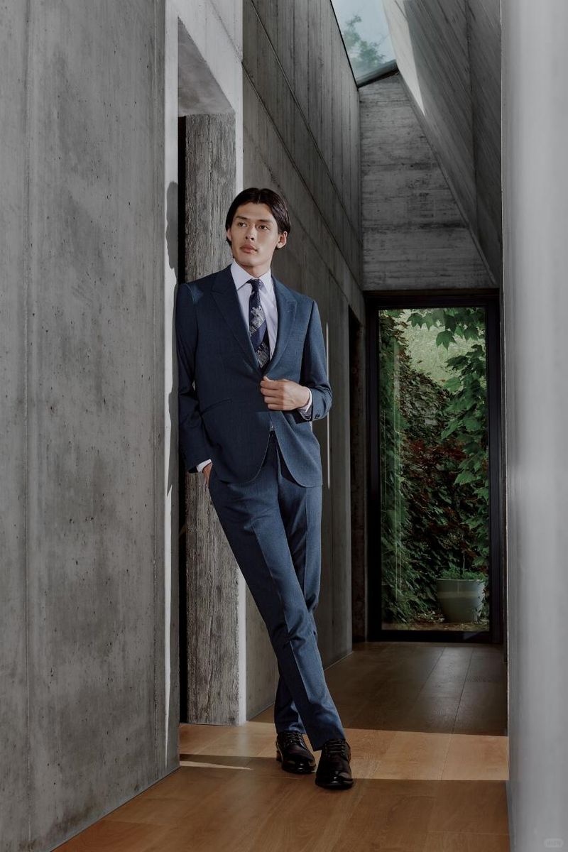 An adult man in a navy suit and black dress shoes poses indoors, leaning against a concrete wall in a brightly lit architectural space.