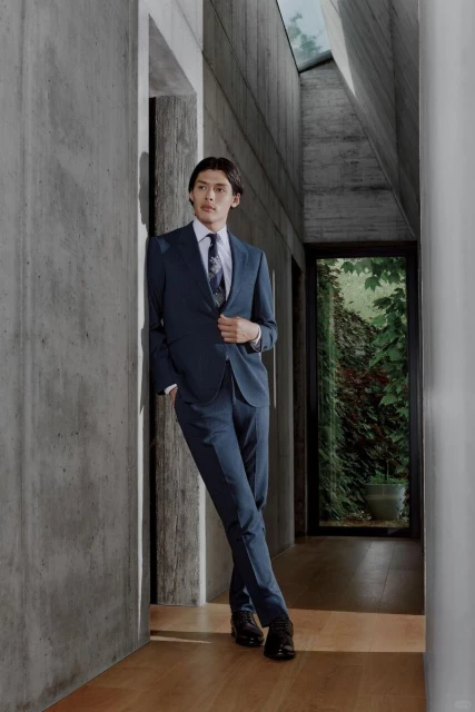 An adult man in a navy suit and black dress shoes poses indoors, leaning against a concrete wall in a brightly lit architectural space.