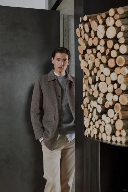 A stylish man stands in a doorway wearing a brown jacket, grey sweater, and tan pants, complemented by a modern interior with a stack of logs.