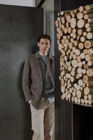 A stylish man stands in a doorway wearing a brown jacket, grey sweater, and tan pants, complemented by a modern interior with a stack of logs.