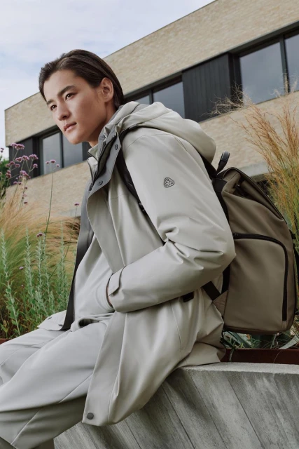 A stylish Asian man in a light-colored jacket and pants sits outdoors with a backpack, set against a modern building and natural grasses.