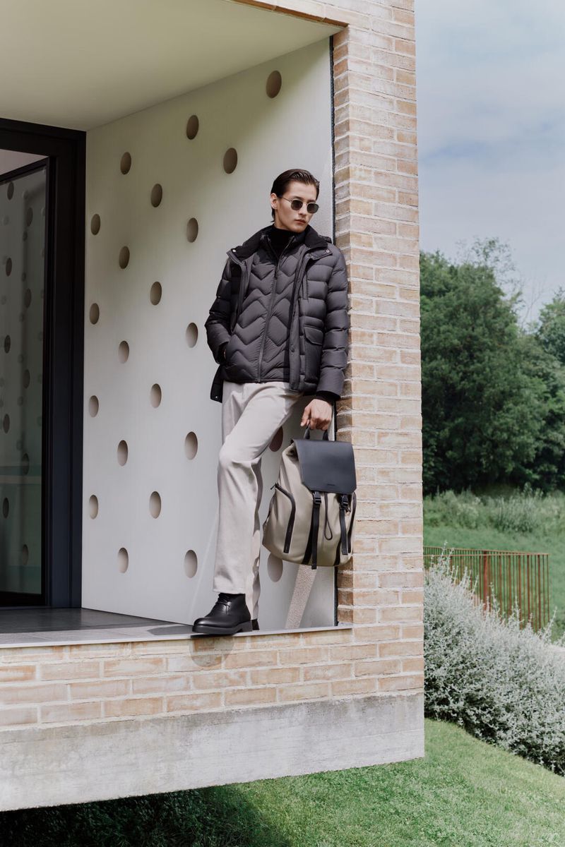 Man wearing a black puffer jacket, light-colored pants, and black boots stands in front of a modern building with a backpack.