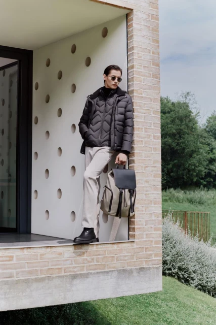 Man wearing a black puffer jacket, light-colored pants, and black boots stands in front of a modern building with a backpack.