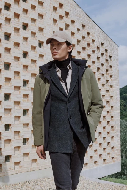 Asian man in a layered outfit featuring a green hooded jacket, blazer, and cap standing outdoors against a modern architectural wall.