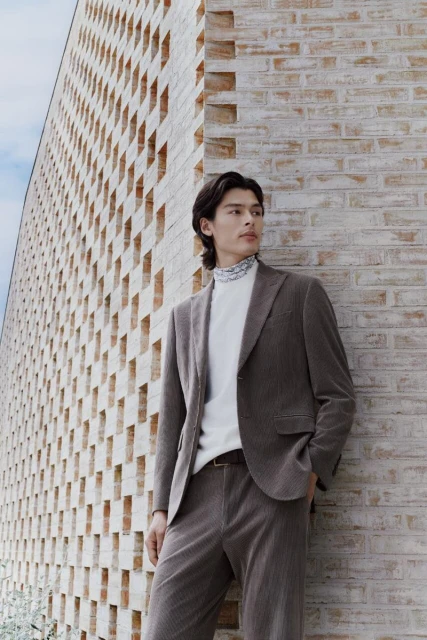 Man in corduroy suit stands against brick wall with cutout pattern. He wears a white turtleneck sweater. Fashion editorial photograph.
