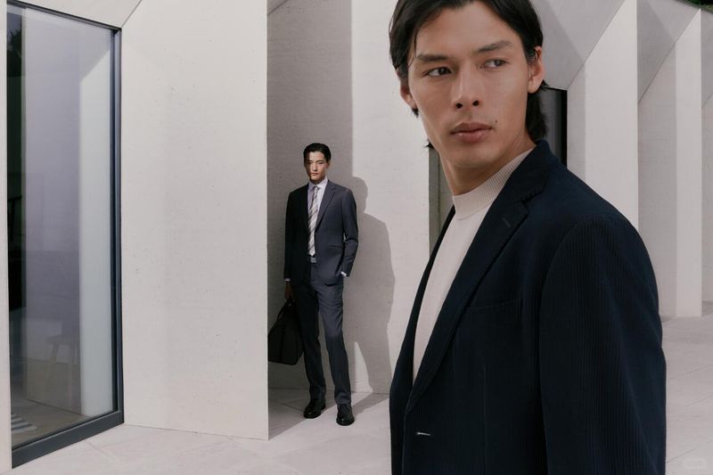 Two adult men in tailored suits pose in a modern architectural setting, one in focus and closer to the camera.