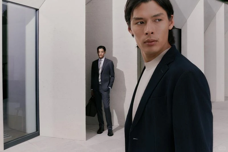 Two adult men in tailored suits pose in a modern architectural setting, one in focus and closer to the camera.