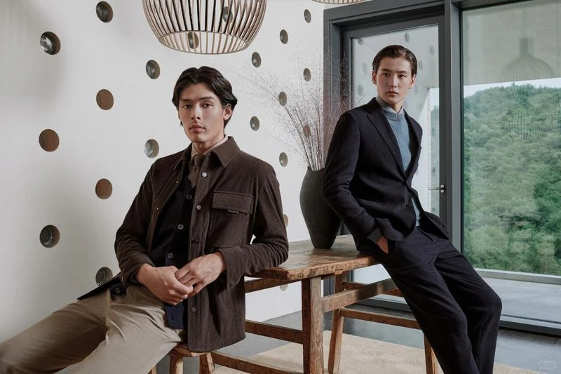 Two stylish young men in jackets and suits pose indoors, leaning against a wooden table in front of a window, with hanging lights above.