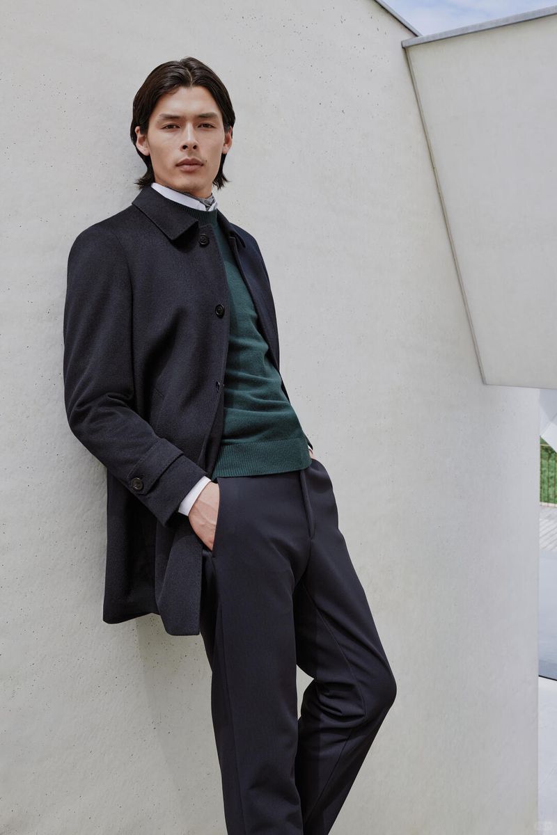 Fashionable man in coat and sweater leaning against modern wall. Sophisticated menswear style. Outdoor fashion shoot with stylish male model.