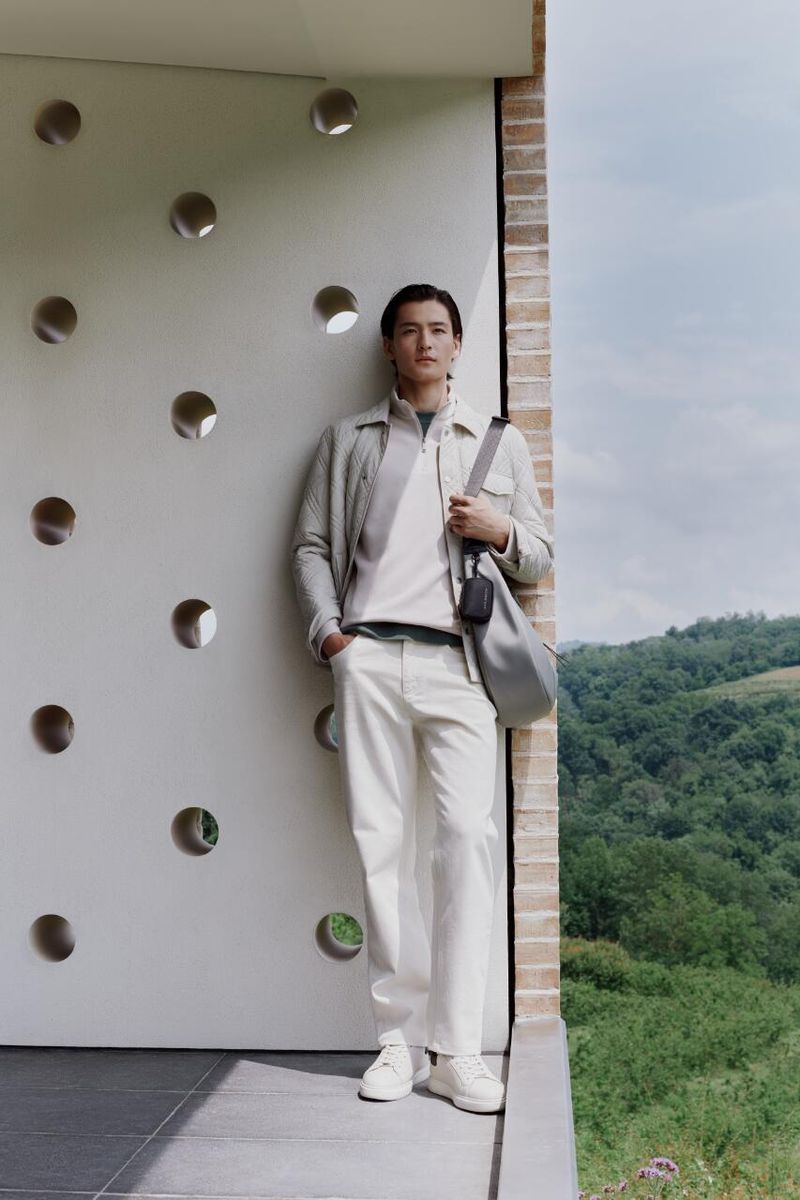 A full-body portrait showcases a male model in an off-white collared outfit, complemented by a shoulder bag, outdoors against a modern building.