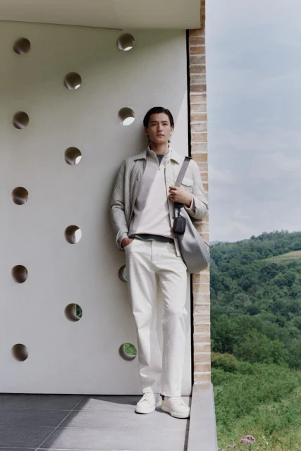 A full-body portrait showcases a male model in an off-white collared outfit, complemented by a shoulder bag, outdoors against a modern building.