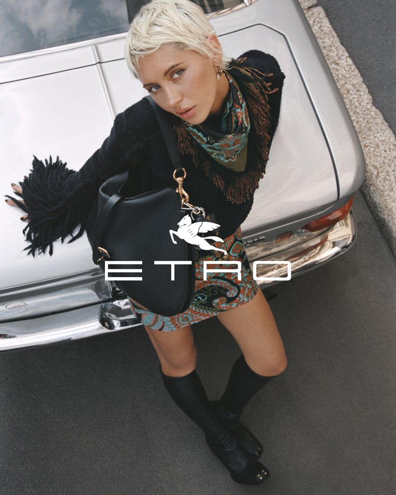 Fashion shot of a woman posing next to a silver car, wearing a patterned mini skirt, a dark sweater, and black boots with an Etro purse.