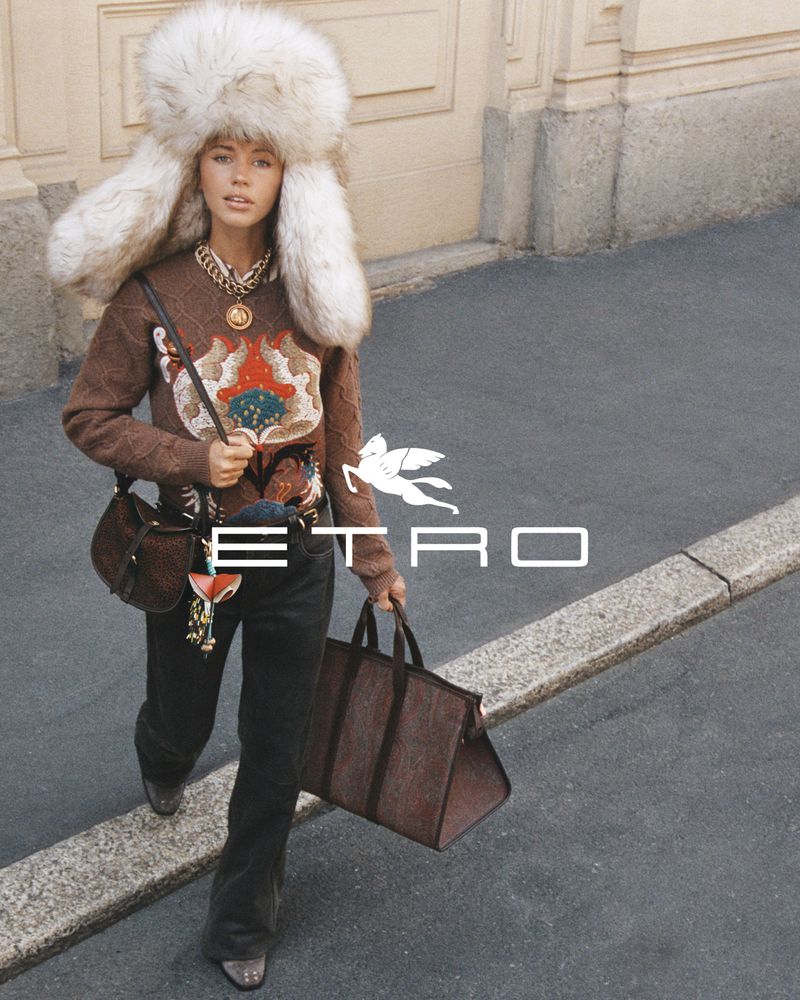 An adult woman walks on a city street wearing a brown sweater, black jeans, fur hat, and holding a brown purse and tote bag.
