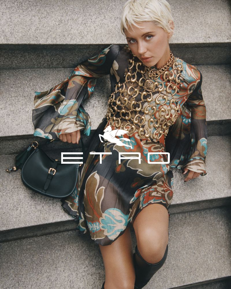 Woman in printed mini dress and gold chain posing on concrete steps with a black shoulder bag for a fashion brand advertisement.