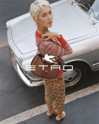 Model Iris Law wearing red patterned sweater and brown leather bag leaning on silver car in Etro campaign.