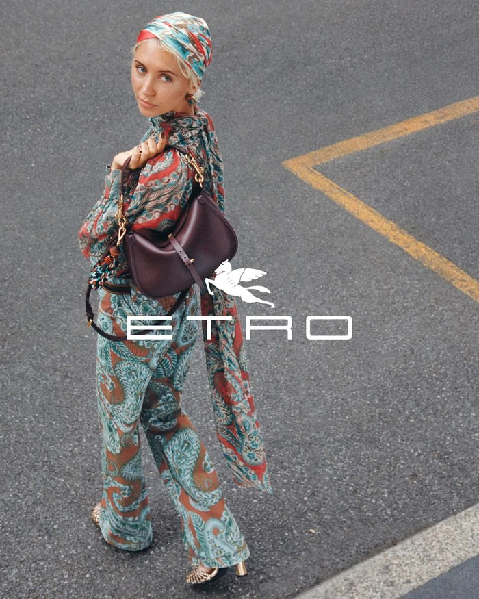 Model Iris Law wearing patterned clothing and a matching headscarf while holding a dark leather shoulder bag on an asphalt street