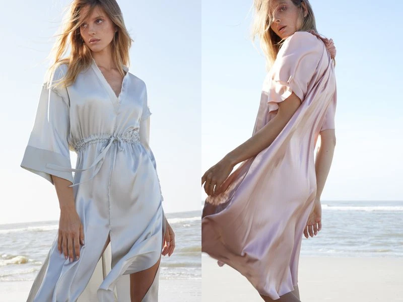 Two women in satin dresses pose on a beach with ocean and blue sky in the background, showcasing fashion and style in outdoor setting.