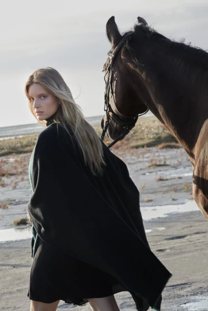 Fashion shot of a blonde woman in a black cape posing next to a brown horse in an open, sandy environment on a cloudy day.