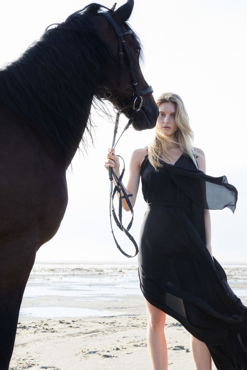 A blond woman in a flowing black dress stands with a dark horse on a sandy beach under a bright sky, creating a serene, elegant scene.