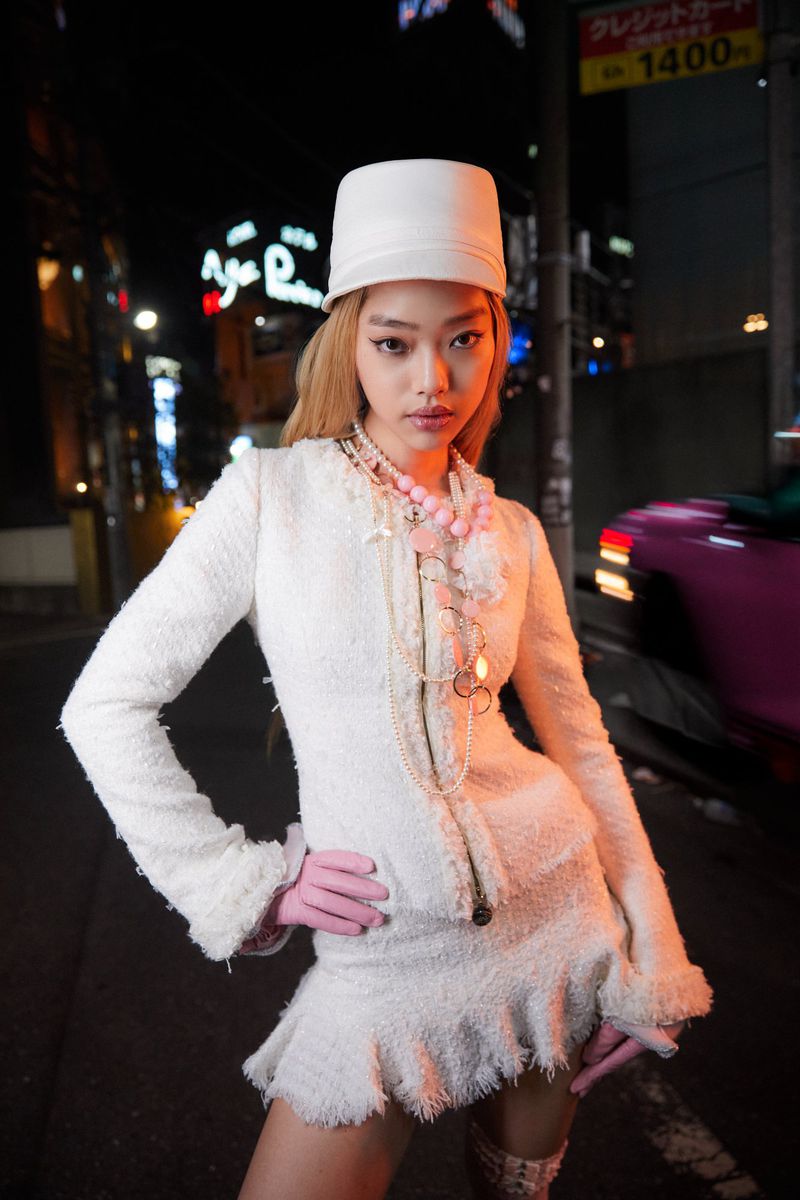 A fashionable young woman in a white textured skirt suit and hat poses on a city street at night, with neon signs in the background.
