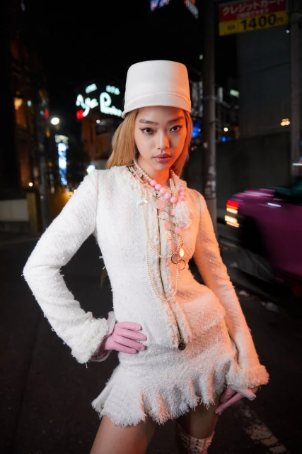 A fashionable young woman in a white textured skirt suit and hat poses on a city street at night, with neon signs in the background.