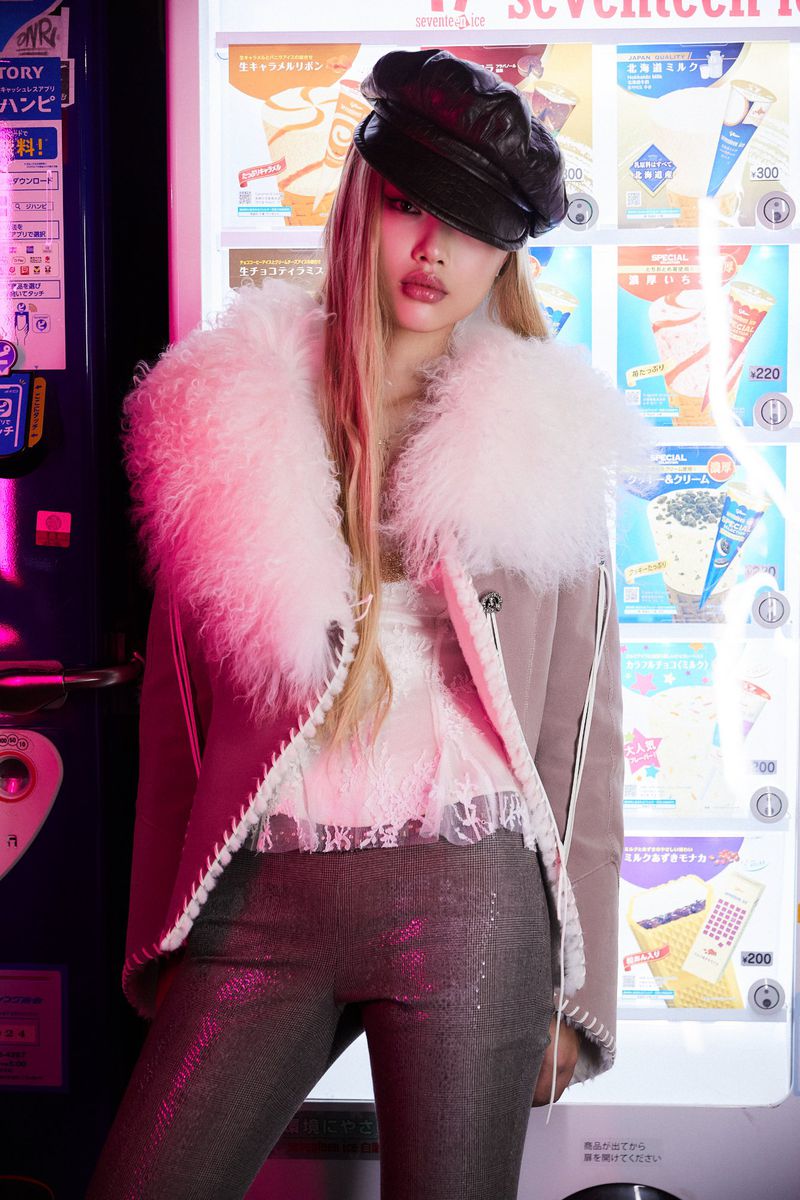 Woman in hat and fur-lined jacket poses in front of a vending machine with product graphics in bright pink and neutral tones.