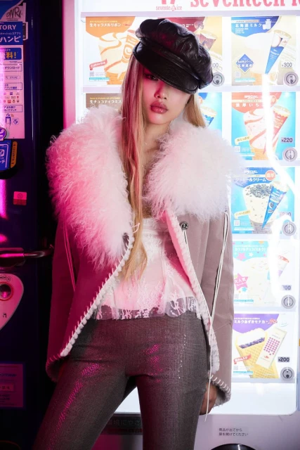 Woman in hat and fur-lined jacket poses in front of a vending machine with product graphics in bright pink and neutral tones.