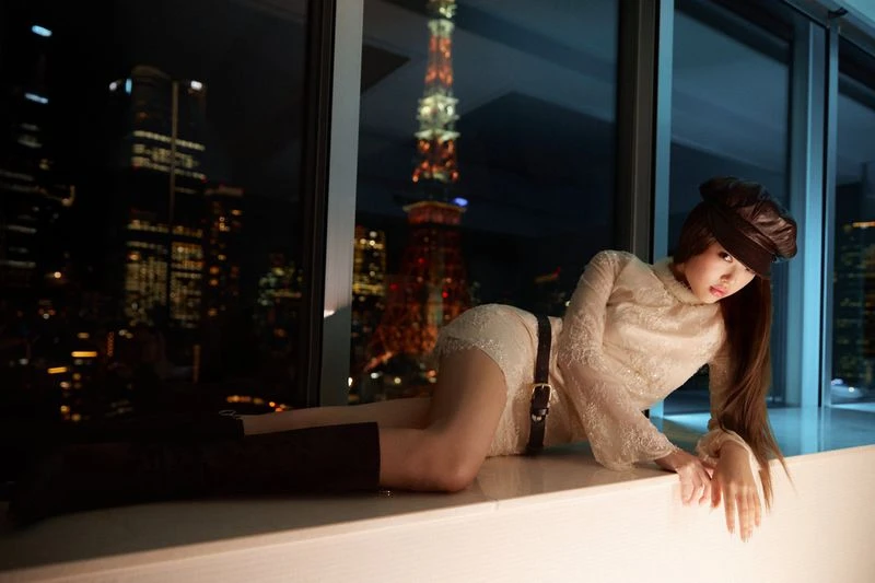 Attractive Asian woman poses in a beige lace dress and brown boots near a window with the Tokyo Tower lit up at night in the background.