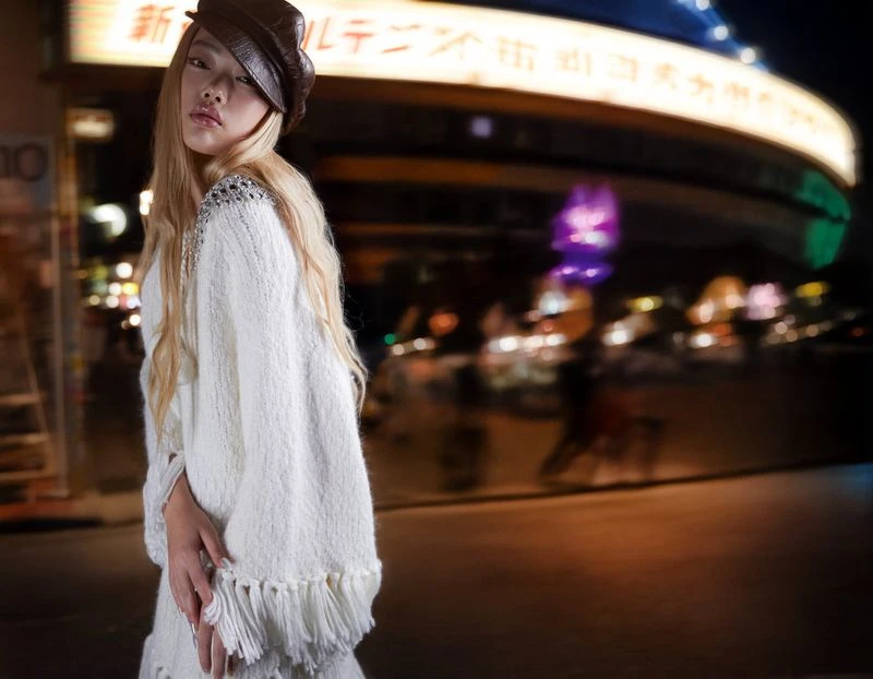 Blonde woman in white fringe sweater and brown cap poses on urban street with blurred lights at night in fashionable outdoor shot.