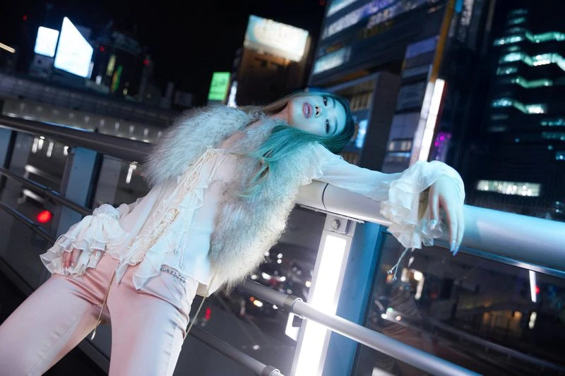 Nighttime fashion shot of a young adult woman with light green hair wearing a white ruffled top and fur vest leaning on a rail in a city.
